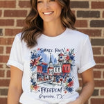 Woman wearing white Small Town Freedom Grapevine TX watercolor patriotic t-shirt at ooh la la! in Grapevine TX 76051