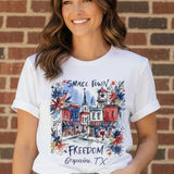 Woman wearing white Small Town Freedom Grapevine TX watercolor patriotic t-shirt at ooh la la! in Grapevine TX 76051