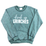 Washed sage green crewneck sweatshirt featuring the white graphic text "drink up" in a script font above the bold block text "GRINCHES." The sweatshirt has a faded, pigment-dyed finish.