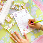 Hand writing scores on a Taylor Paladino Mahjong Scorecard Pad surrounded by decorative tiles