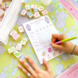 Hand writing scores on a Taylor Paladino Mahjong Scorecard Pad surrounded by decorative tiles